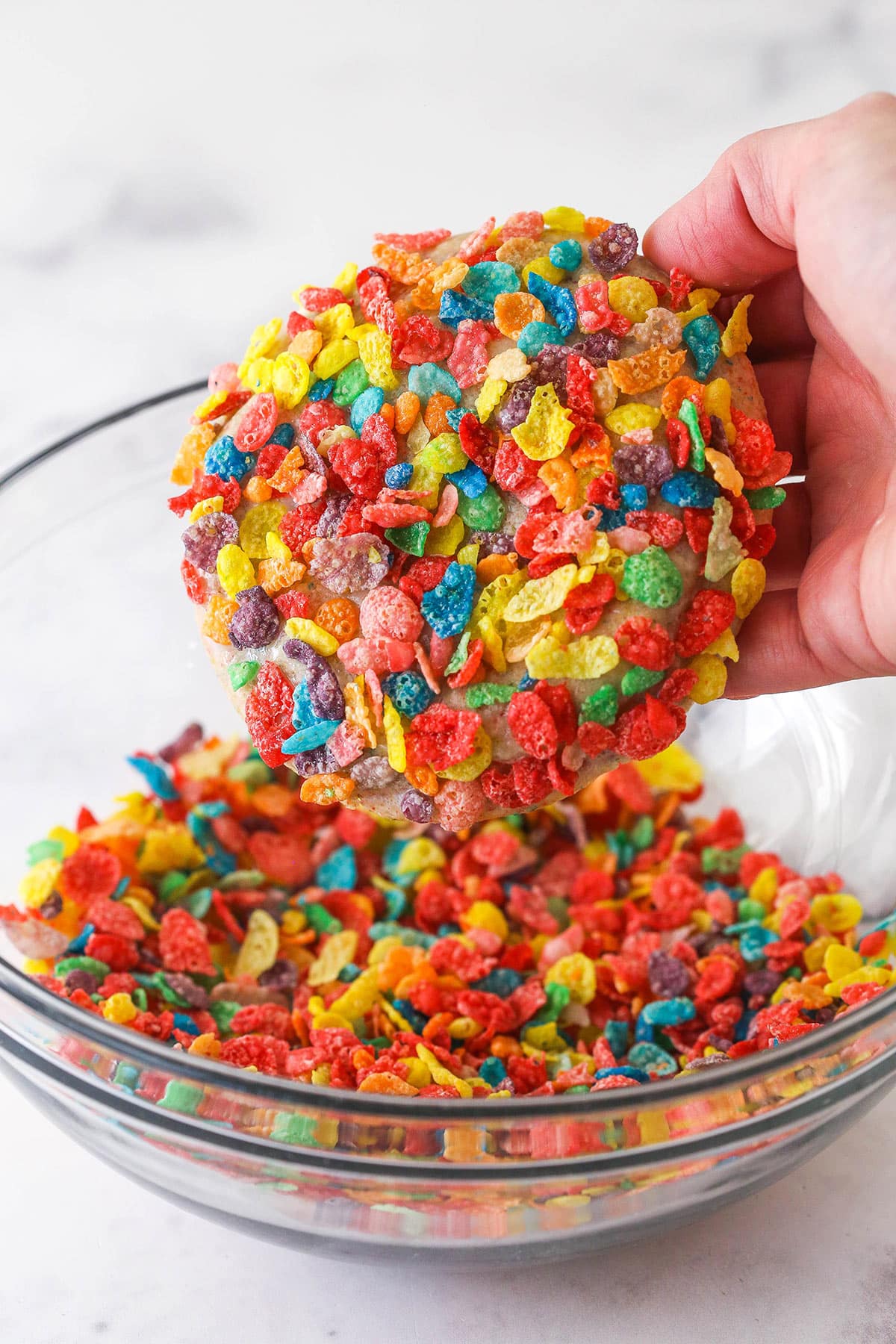 Chewy breakfast cereal infused cookies are coated in a light glaze and dipped in bright Fruity Pebbles to make rainbow Fruity Pebbles Cookies. Coating a Fruity Pebbles cookie in Fruity Pebbles in a bowl.
