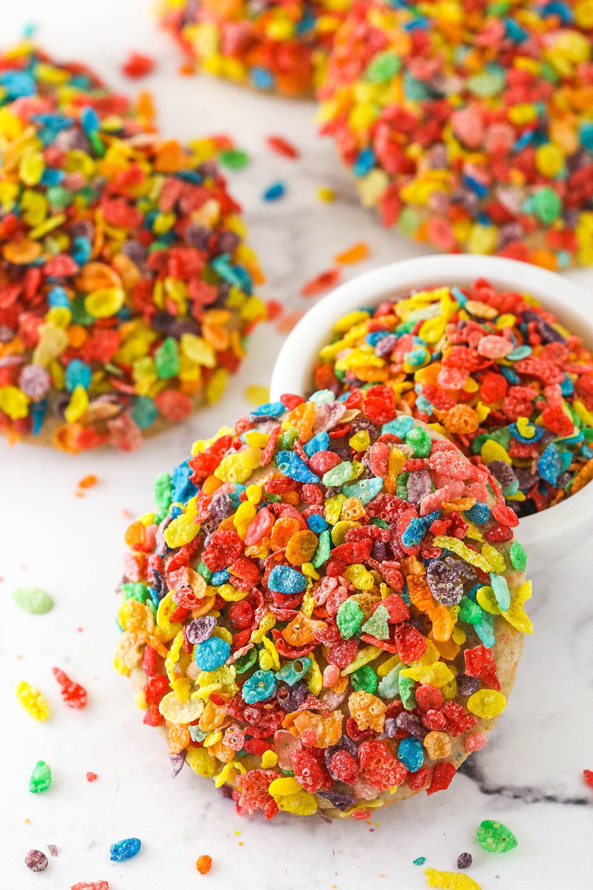 A bowl of Fruit Pebbles with Fruity Pebbles cookie leaning against it surrounded by more Fruity Pebbles cookies.