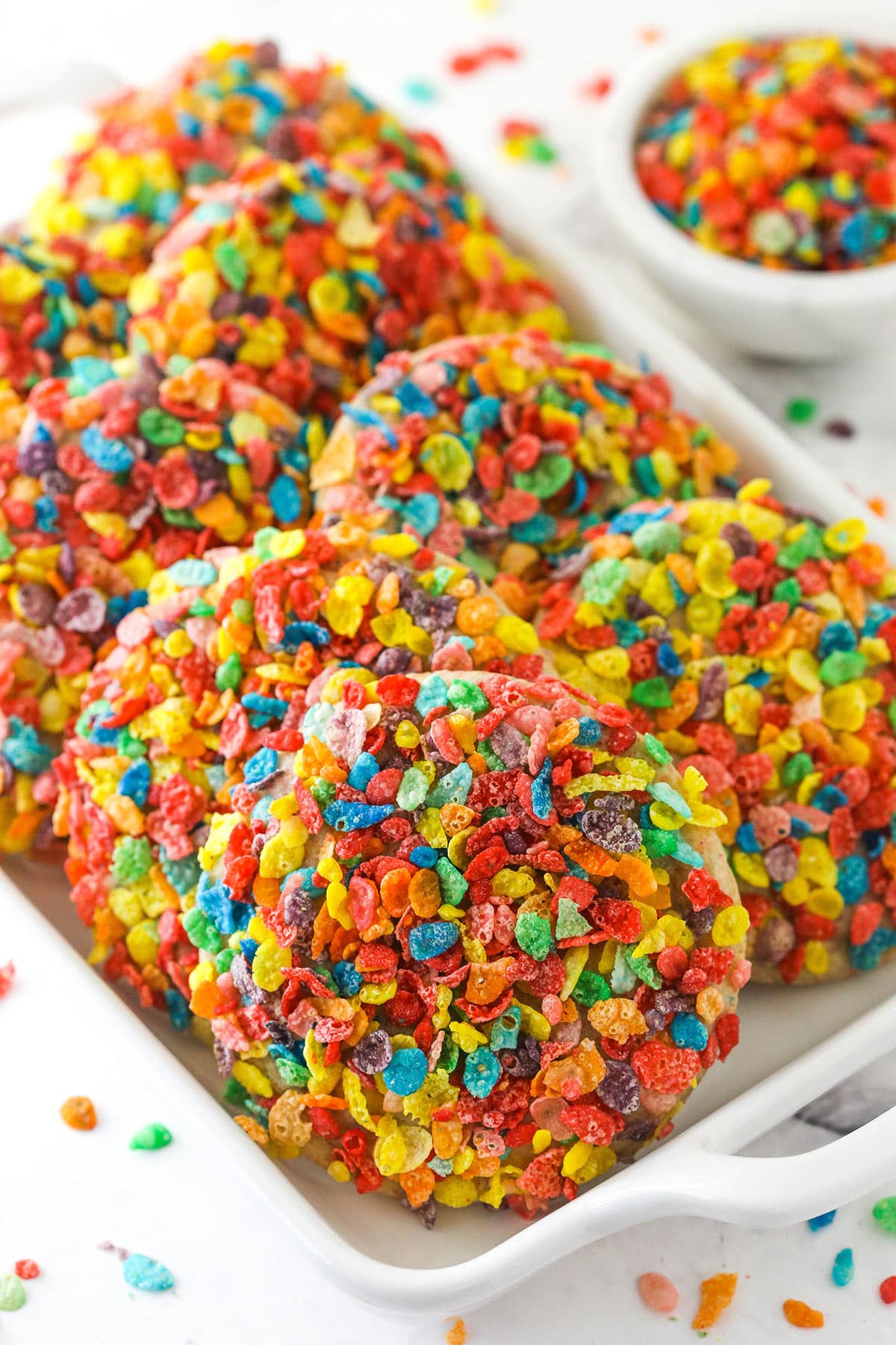 Fruity Pebbles Cookies arrange on a serving platter near a bowl of Fruity Pebbles.