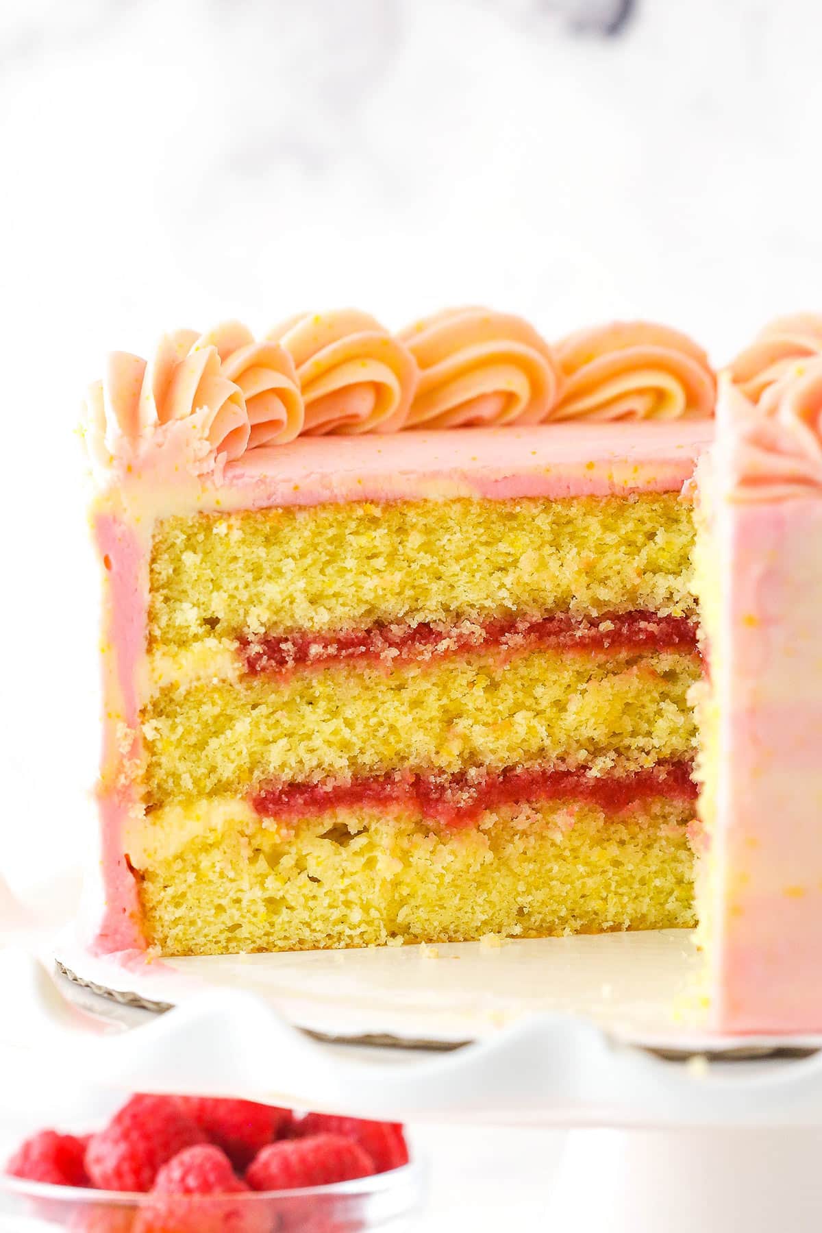 Tender orange cake, raspberry filling, cream cheese frosting and a drizzle of orange syrup come together in this raspberry orange layer cake. Raspberry orange layer cake on a cake stand with a slice cut out of it near a bowl of raspberries.