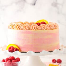 Raspberry orange layer cake on a cake stand with fresh raspberries and orange slices nearby.