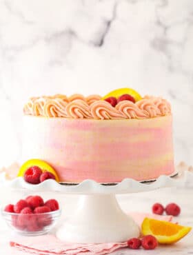 Raspberry orange layer cake on a cake stand with fresh raspberries and orange slices nearby.