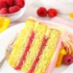 A slice of raspberry orange layer cake on a plate with a fork surrounded by fresh raspberries and orange slices.