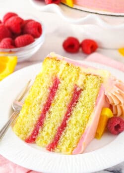 A slice of raspberry orange layer cake on a plate with a fork surrounded by fresh raspberries and orange slices.