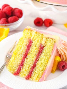 A slice of raspberry orange layer cake on a plate with a fork surrounded by fresh raspberries and orange slices.