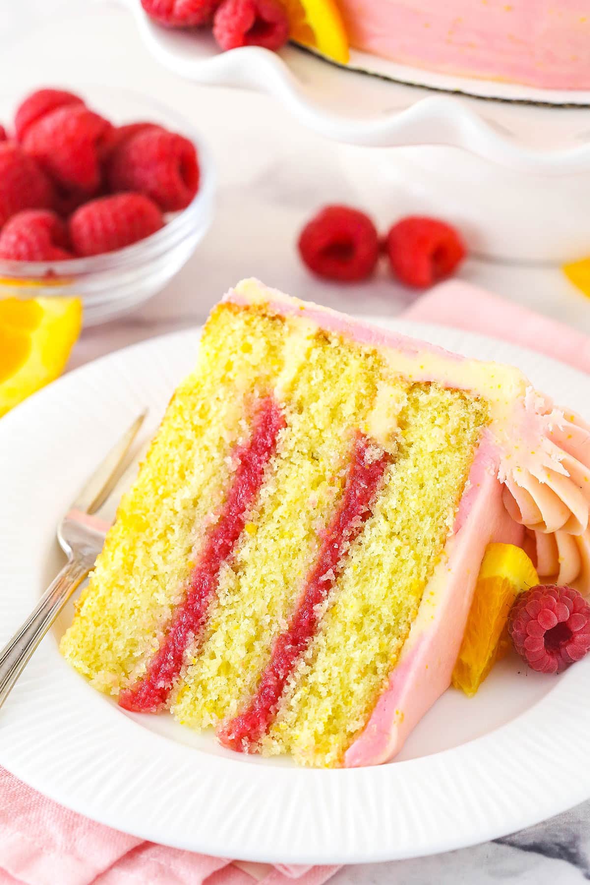 A slice of raspberry orange layer cake on a plate with a fork surrounded by fresh raspberries and orange slices.