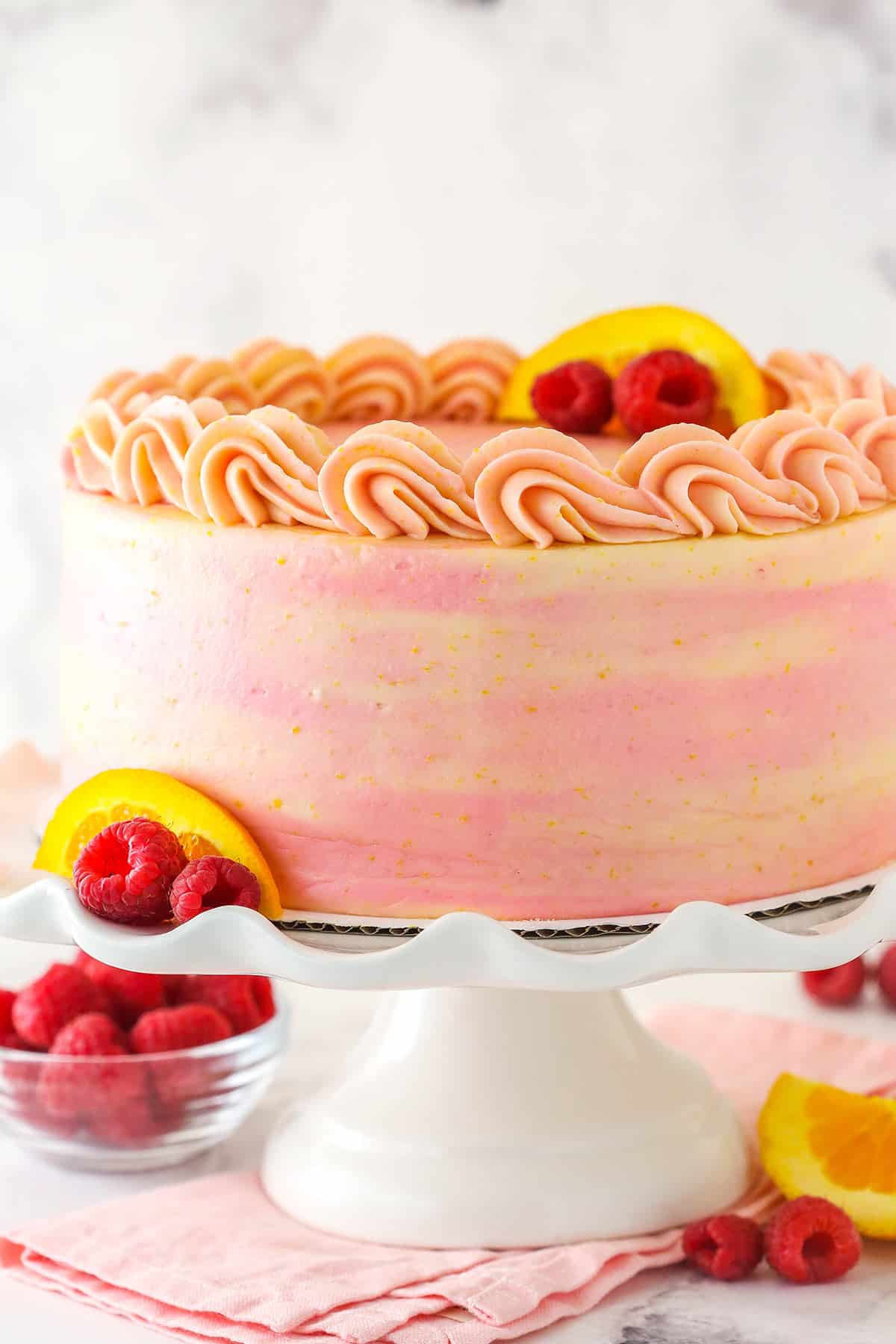 Tender orange cake, raspberry filling, cream cheese frosting and a drizzle of orange syrup come together in this raspberry orange layer cake. Raspberry orange layer cake on a cake stand with fresh raspberries and orange slices nearby.