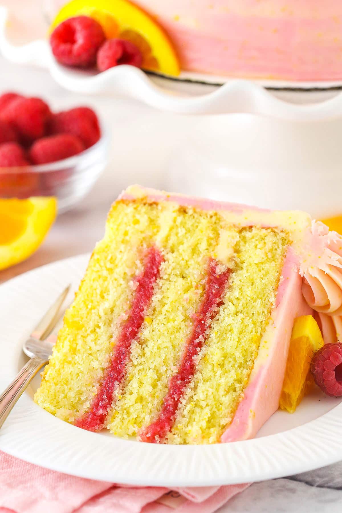 A slice of raspberry orange layer cake on a plate with a fork surrounded by fresh raspberries and orange slices.