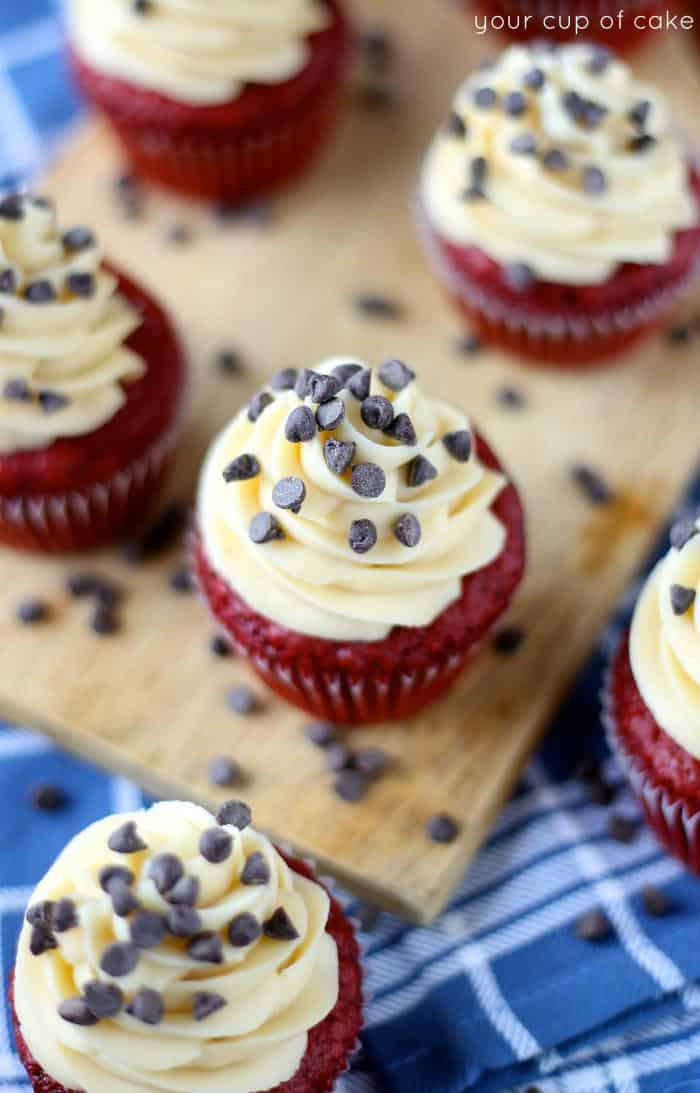 Several red velvet cookie dough cupcakes on a cutting board surrounded by mini chocolate chips. 