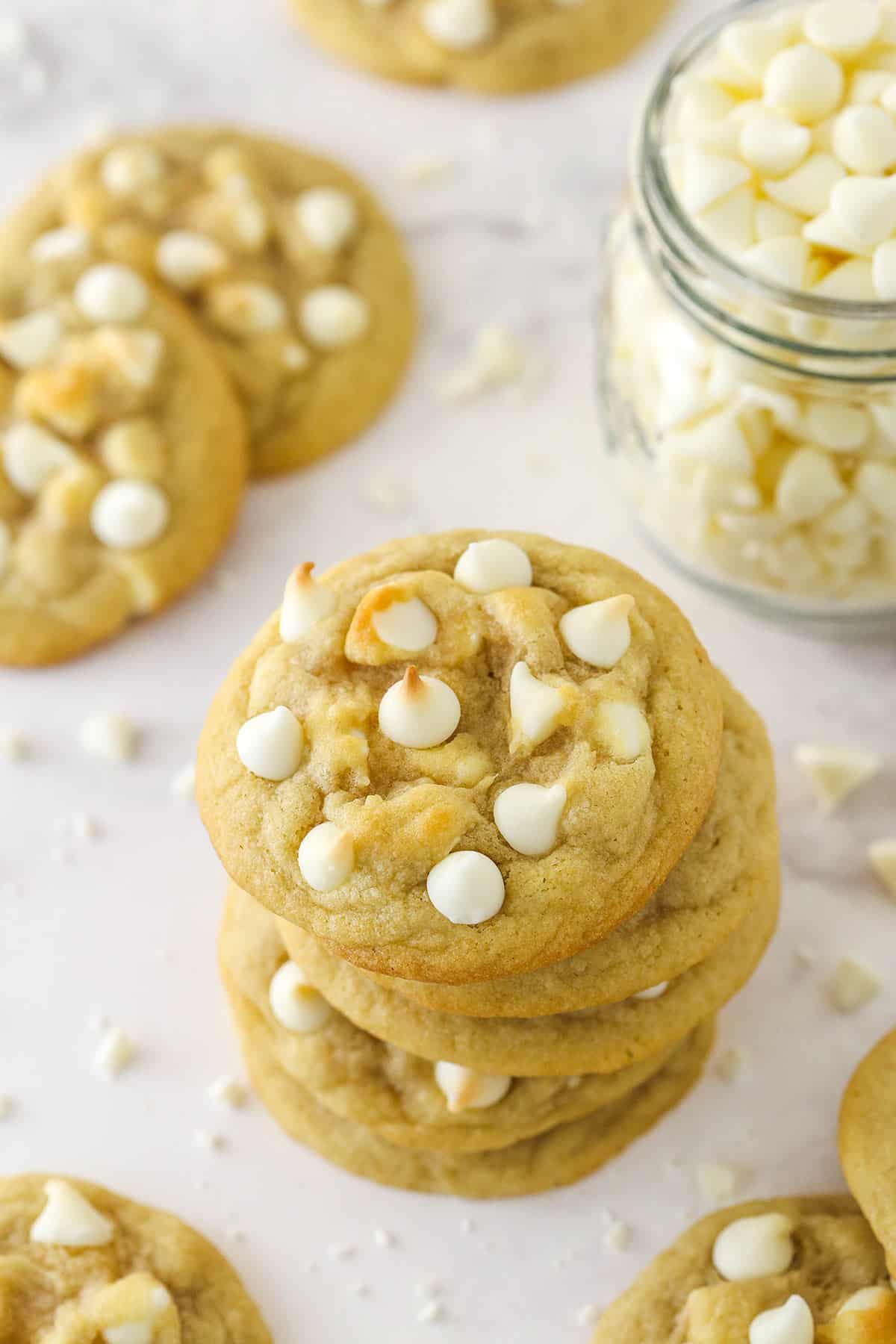 A stack of white chocolate chip cookies surrounded by other white chocolate chip cookies near a jar of white chocolate chips.