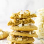 A stack of white chocolate chip cookies surrounded by other white chocolate chip cookies and with the top cookie broken in half.