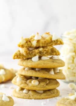 A stack of white chocolate chip cookies surrounded by other white chocolate chip cookies and with the top cookie broken in half.