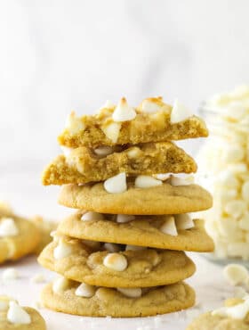 A stack of white chocolate chip cookies surrounded by other white chocolate chip cookies and with the top cookie broken in half.