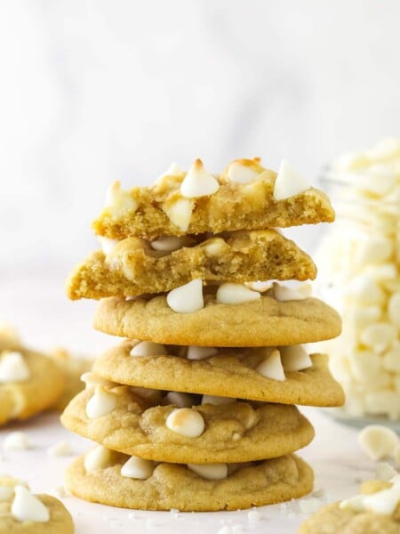 A stack of white chocolate chip cookies surrounded by other white chocolate chip cookies and with the top cookie broken in half.