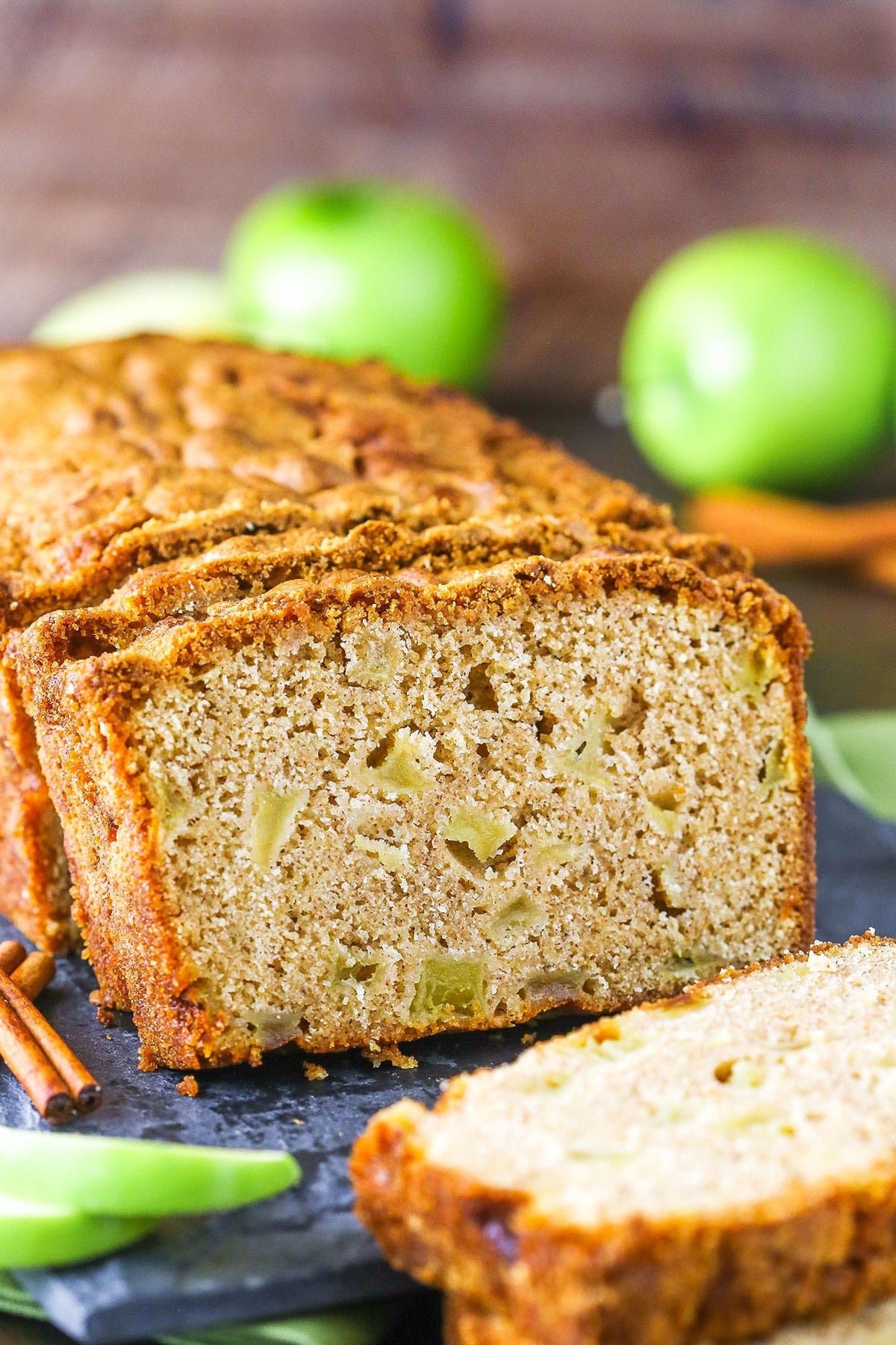 Filled with warm cinnamon and fresh apples, this homemade Apple Bread is the best Fall breakfast or snack recipe! It's so soft, moist and dense with a perfectly crispy crust. Side view of a loaf of Apple Bread with the end slice removed on a black cutting board with green apples in the background