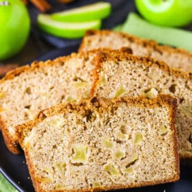 Four slices of Apple Bread on a black plate with whole and cut apples in the background