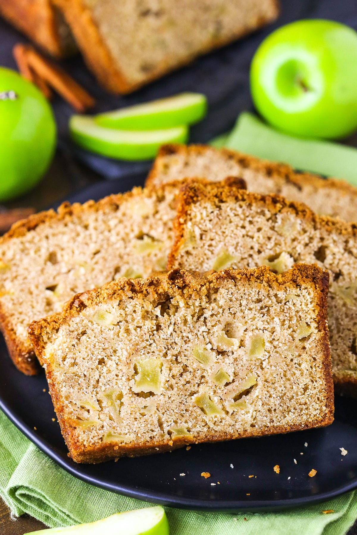 Four slices of Apple Bread on a black plate with whole and cut apples in the background