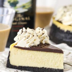 A slice of Baileys cheesecake on a plate near a bottle of Baileys Irish Cream and 2 glasses of Baileys Irish Cream.