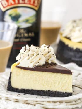 A slice of Baileys cheesecake on a plate near a bottle of Baileys Irish Cream and 2 glasses of Baileys Irish Cream.