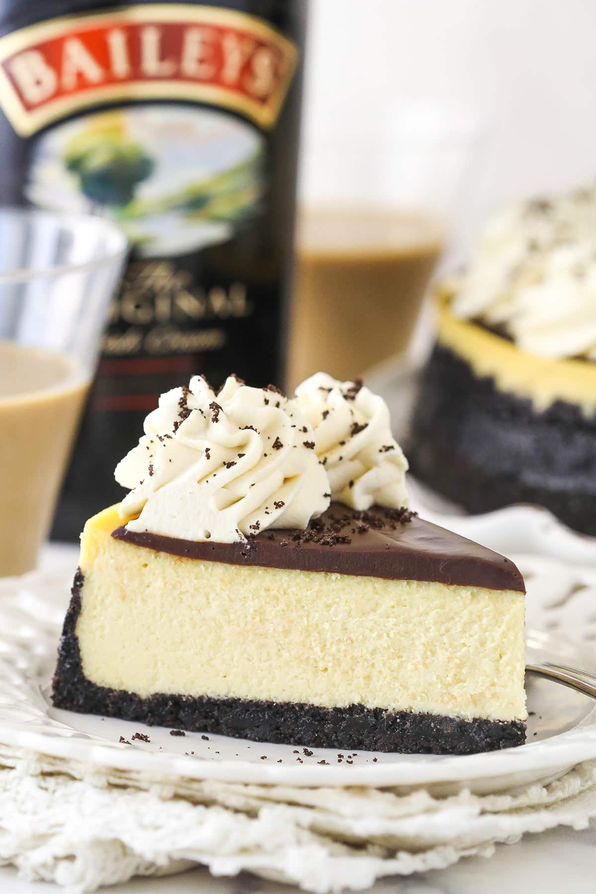 Oreo crust, creamy Baileys cheesecake filling, Baileys chocolate ganache, and Baileys whipped cream come together in this Baileys cheesecake. A slice of Baileys cheesecake on a plate near a bottle of Baileys Irish Cream and 2 glasses of Baileys Irish Cream.
