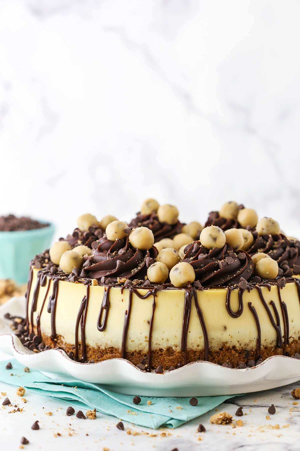 This Chocolate Chip Cookie Dough Cheesecake is made with a chocolate chip cookie crust and brown sugar cheesecake filled with chocolate chips and cookie dough! It's topped with chocolate ganache and more cookie dough for the ultimate treat! side image of cookie dough cheesecake with blue bowl in background