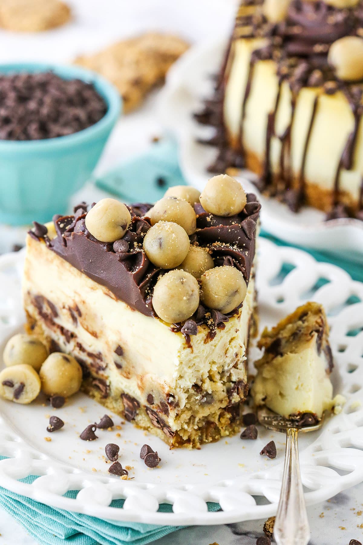 This Chocolate Chip Cookie Dough Cheesecake is made with a chocolate chip cookie crust and brown sugar cheesecake filled with chocolate chips and cookie dough! It's topped with chocolate ganache and more cookie dough for the ultimate treat! cheesecake with a bite taken, teal bowl in background