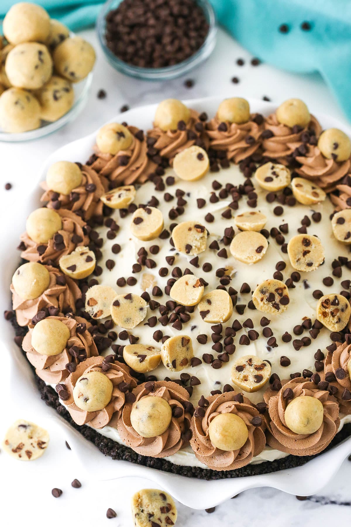 Overhead view of a full Chocolate Chip Cookie Dough Ice Cream Pie in a white platter with cookie dough balls and chocolate chips in the background