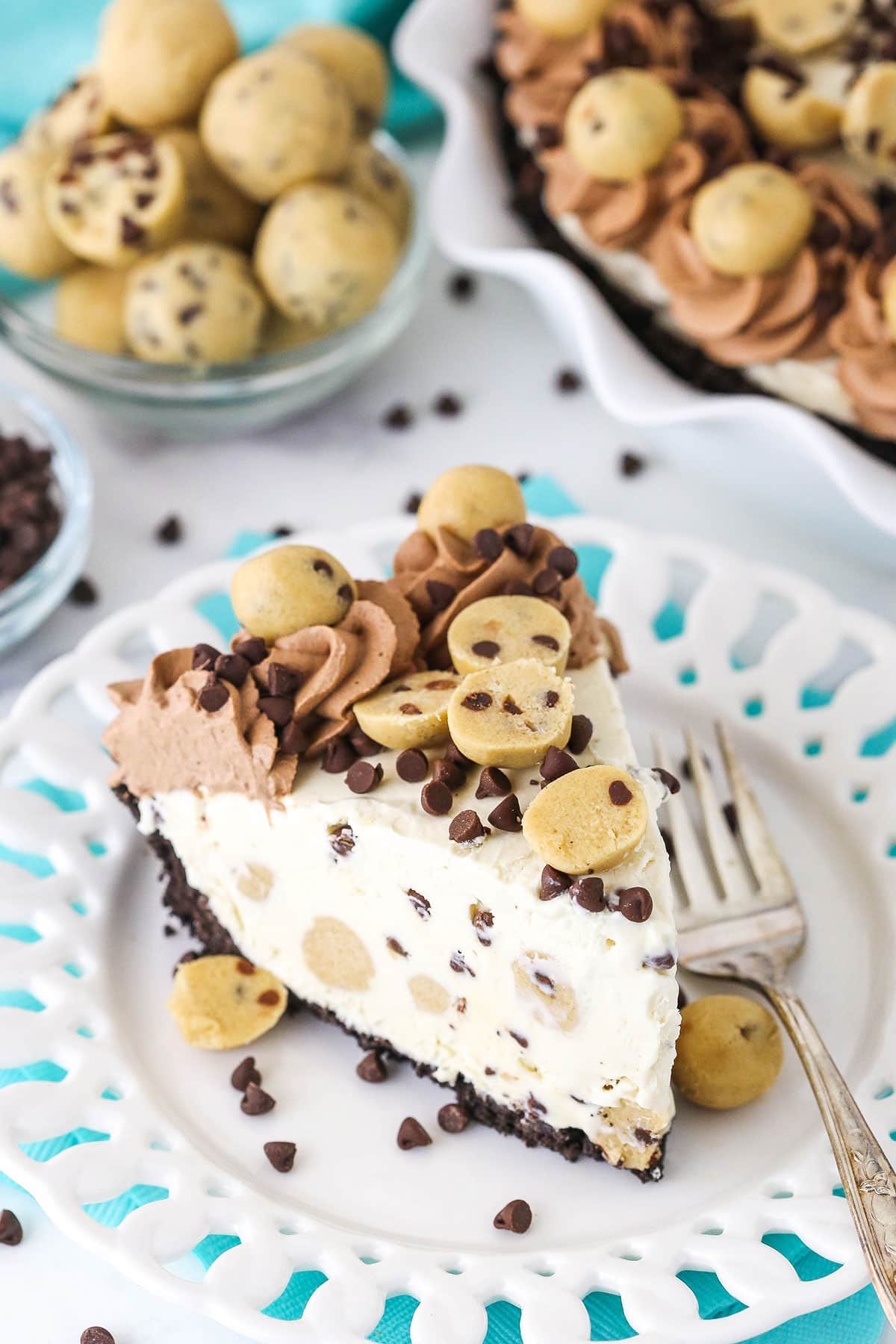 Chocolate Chip Cookie Dough Ice Cream Pie is oh so delicious! This easy no bake dessert has an Oreo crust, no churn vanilla ice cream filled with edible cookie dough and mini chocolate chips! Slice of Chocolate Chip Cookie Dough Ice Cream Pie on a white plate with a fork and cookie dough balls and chocolate chips in the background