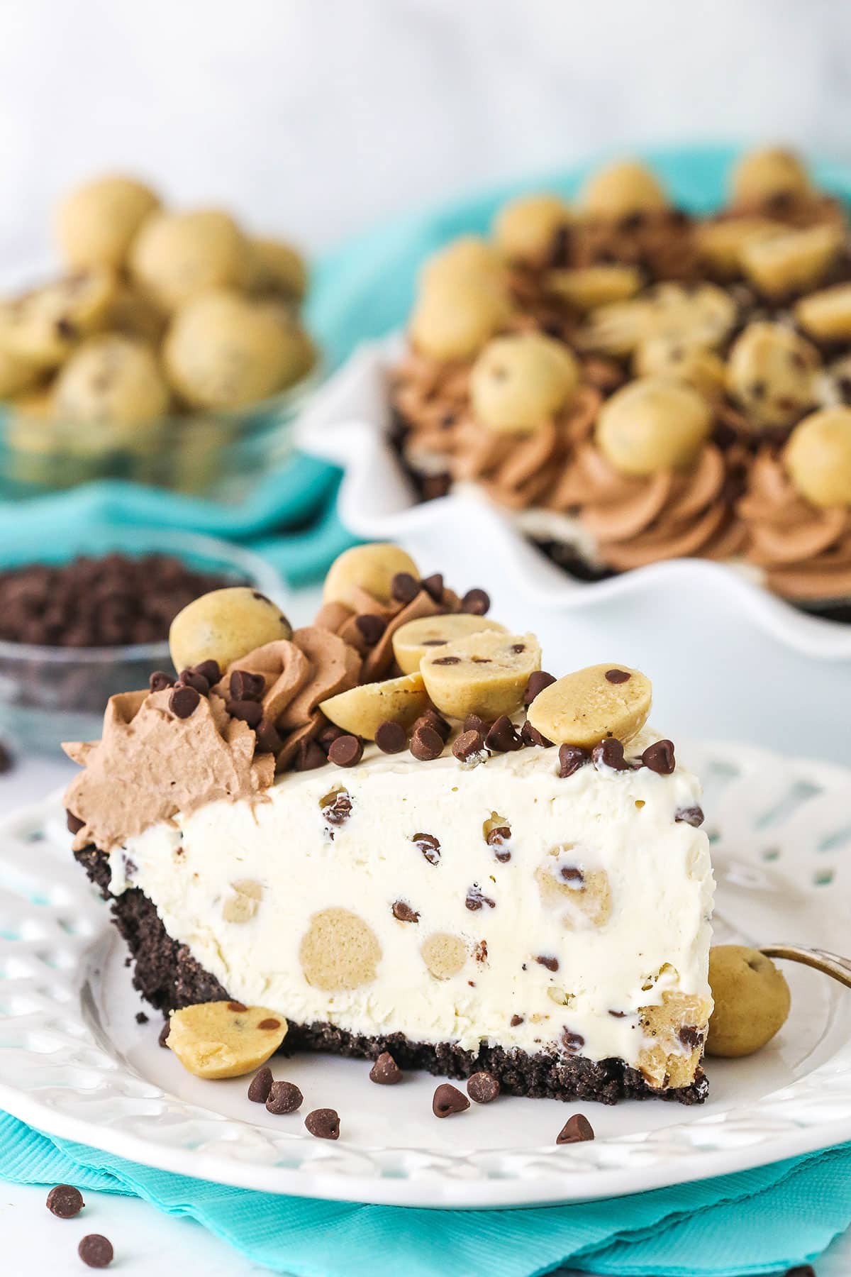 Slice of Chocolate Chip Cookie Dough Ice Cream Pie on a white plate with a fork and cookie dough balls and chocolate chips in the background