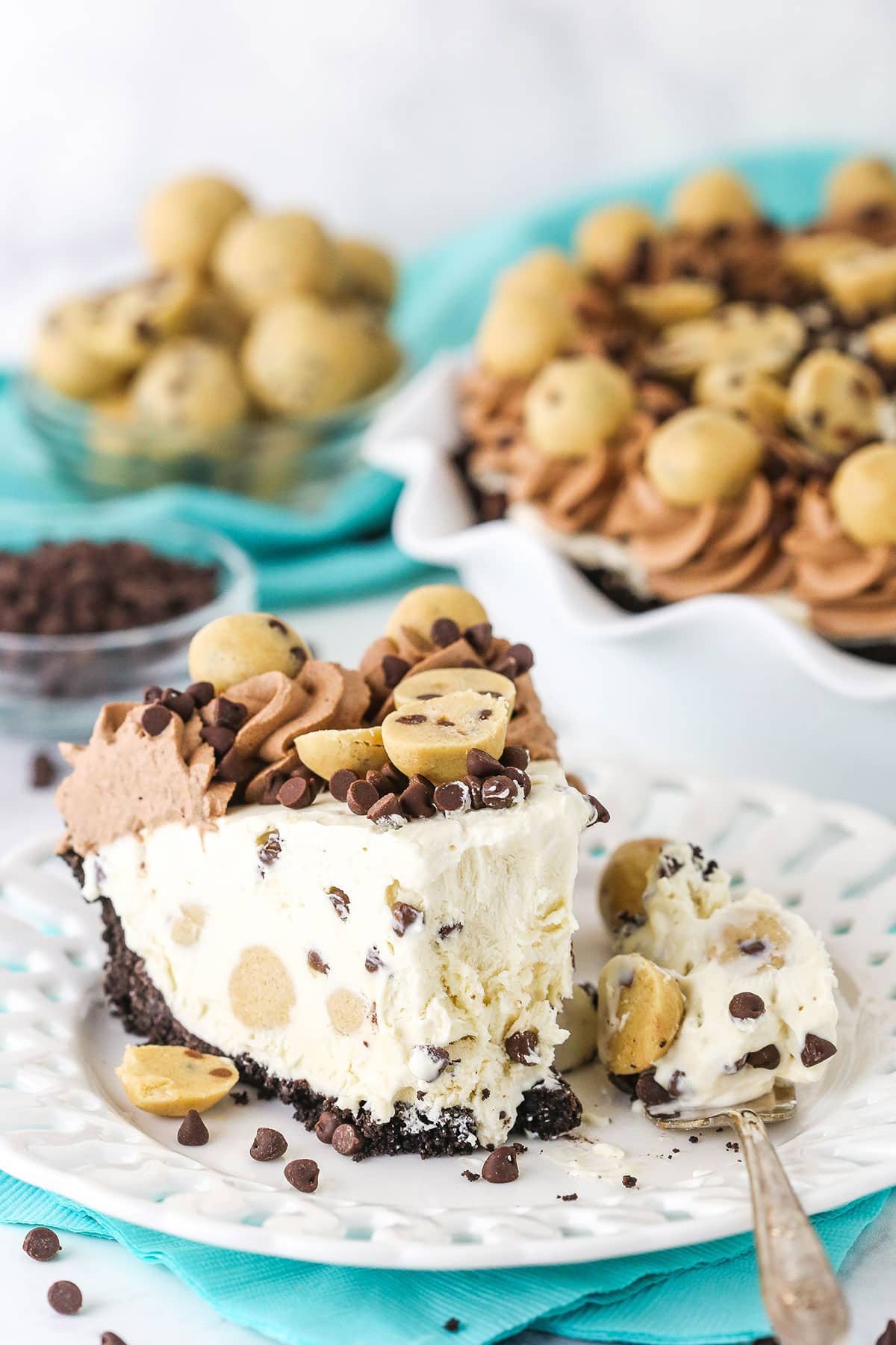 Slice of Chocolate Chip Cookie Dough Ice Cream Pie with a bite taken out on a white plate with a fork and cookie dough balls and chocolate chips in the background