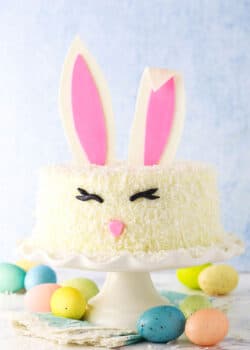 Side shot of a complete Easter Bunny Cake with fondant bunny ears, eyes and nose on a white cake stand with painted Easter eggs underneath