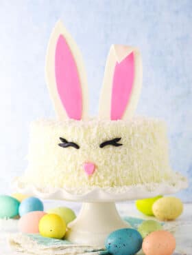Side shot of a complete Easter Bunny Cake with fondant bunny ears, eyes and nose on a white cake stand with painted Easter eggs underneath