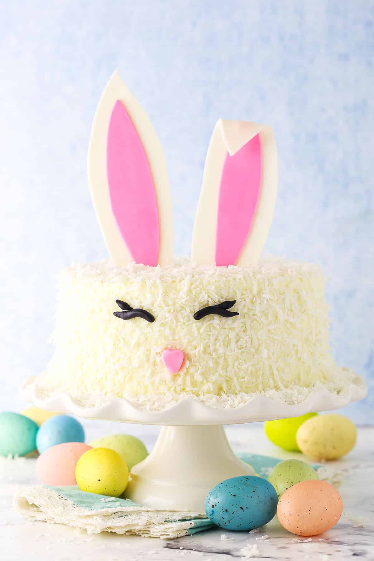 Side shot of a complete Easter Bunny Cake with fondant bunny ears, eyes and nose on a white cake stand with painted Easter eggs underneath