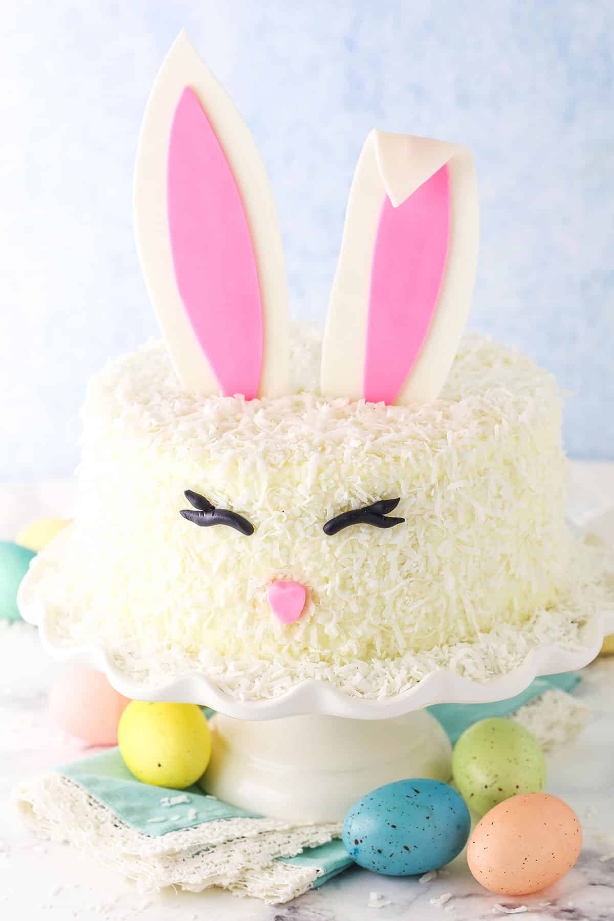 Side shot of a complete Easter Bunny Cake with fondant bunny ears, eyes and nose on a white cake stand with painted Easter eggs underneath