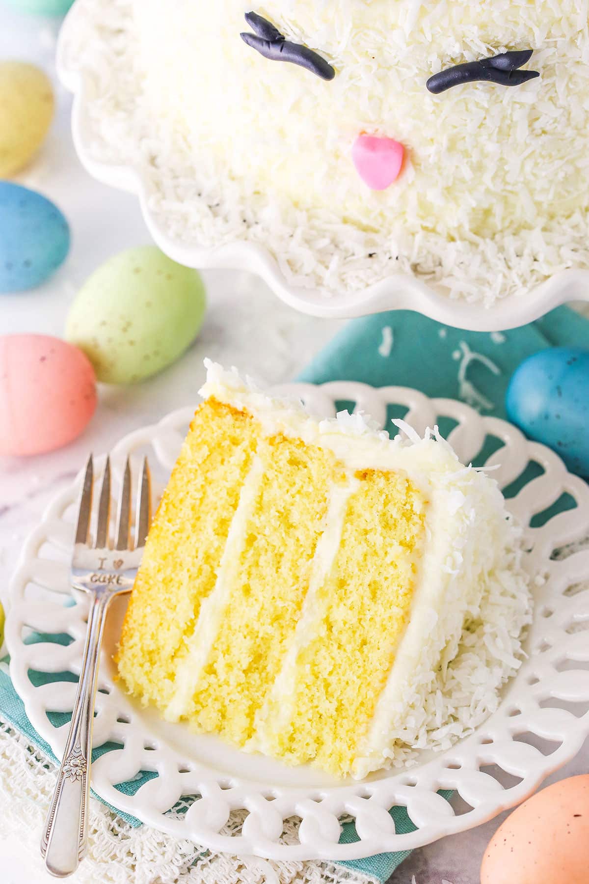 Overhead view of a slice of Easter Bunny Cake on a white plate with a fork with painted Easter eggs surrounding