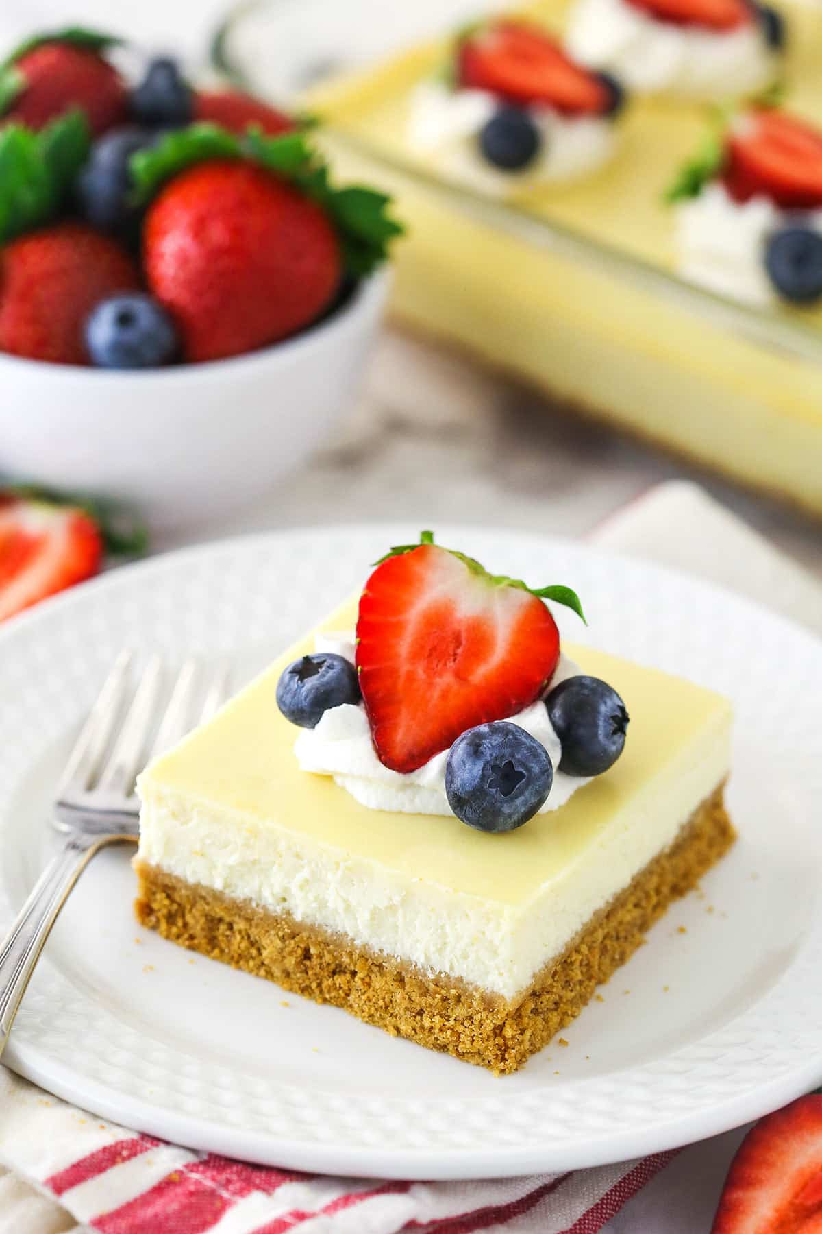 A slice of easy cheesecake on a plate with a fork near a bowl of berries and a full cheesecake.