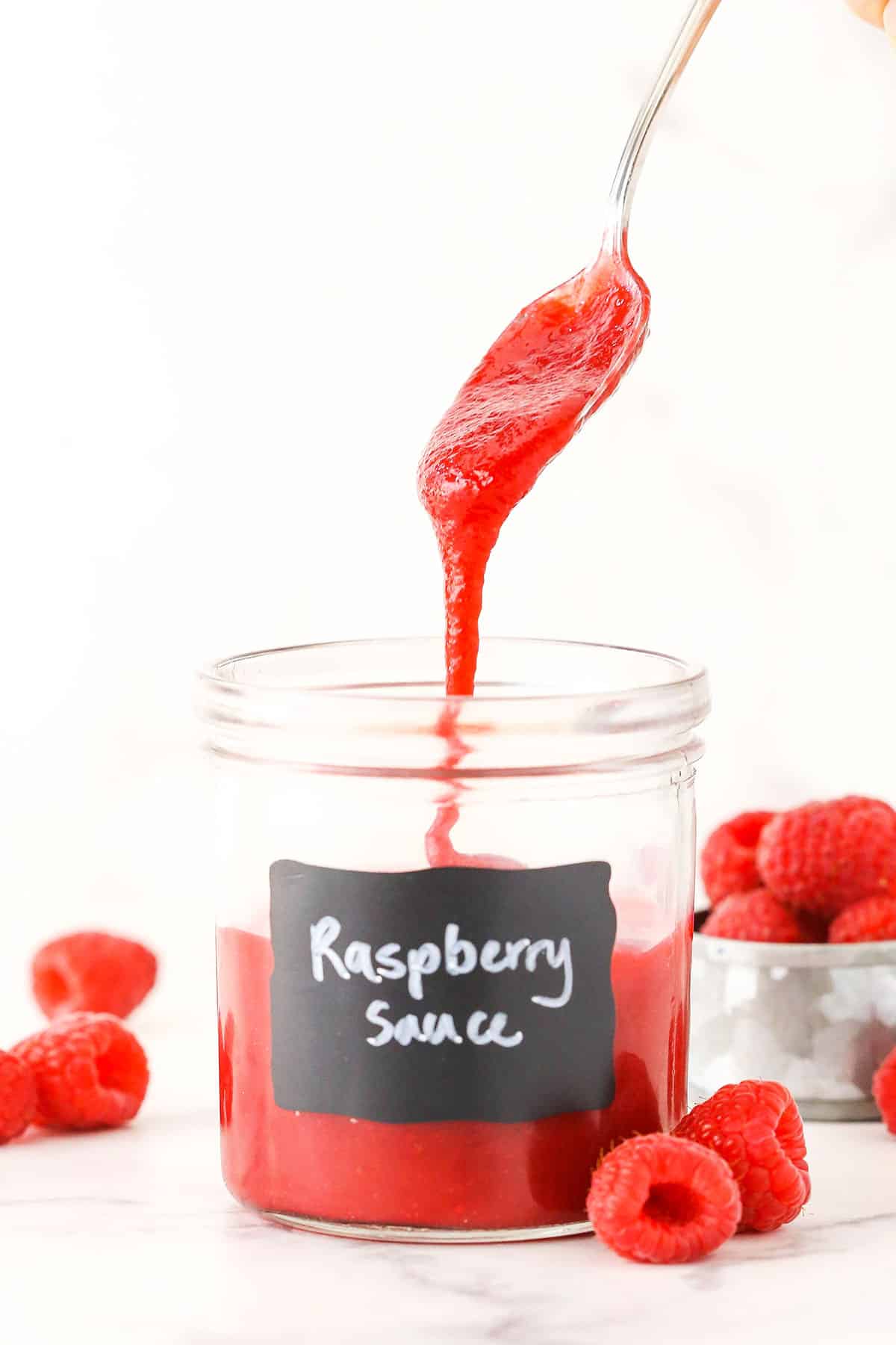 Raspberry sauce dripping off a spoon into a clear glass jar with raspberries in the background