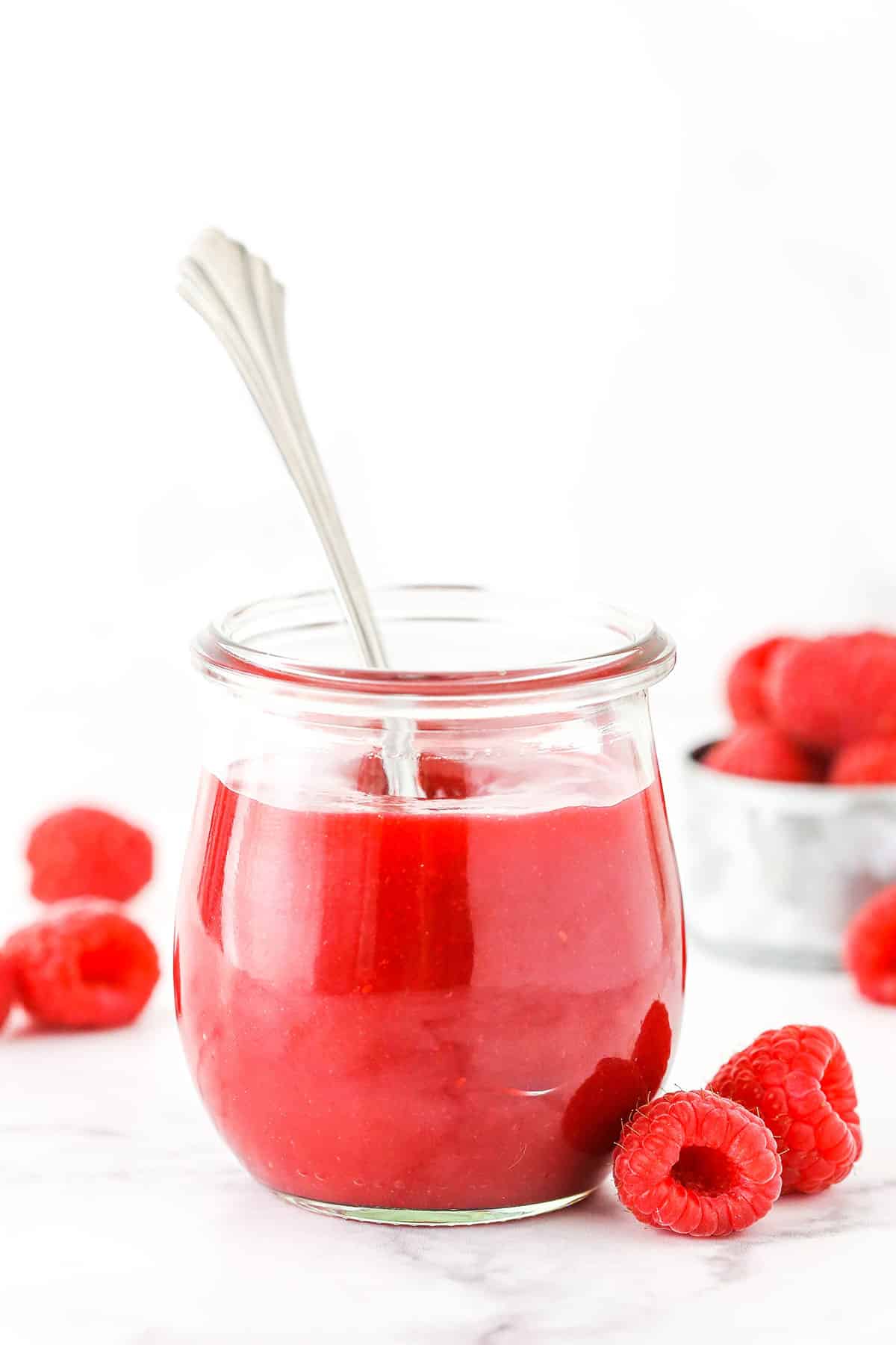 Raspberry sauce in a clear glass jar with a spoon and raspberries in the background