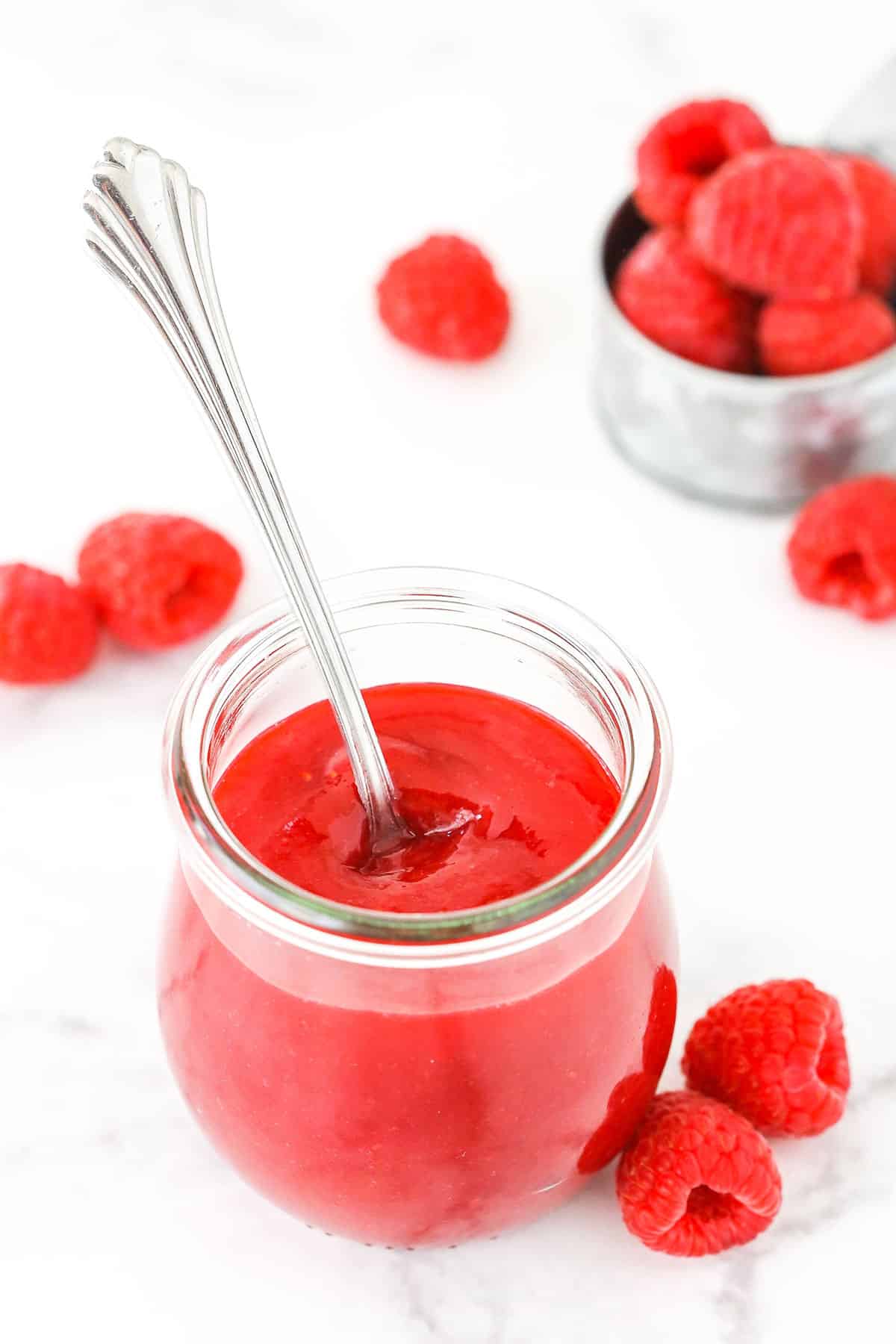 Raspberry sauce in a clear glass jar with a spoon and raspberries in the background