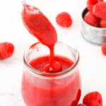 Raspberry sauce dripping off a spoon into a clear glass jar with raspberries in the background