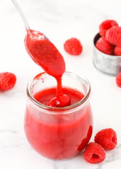 Raspberry sauce dripping off a spoon into a clear glass jar with raspberries in the background