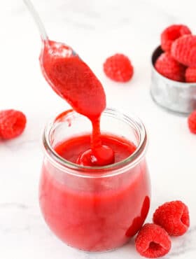 Raspberry sauce dripping off a spoon into a clear glass jar with raspberries in the background