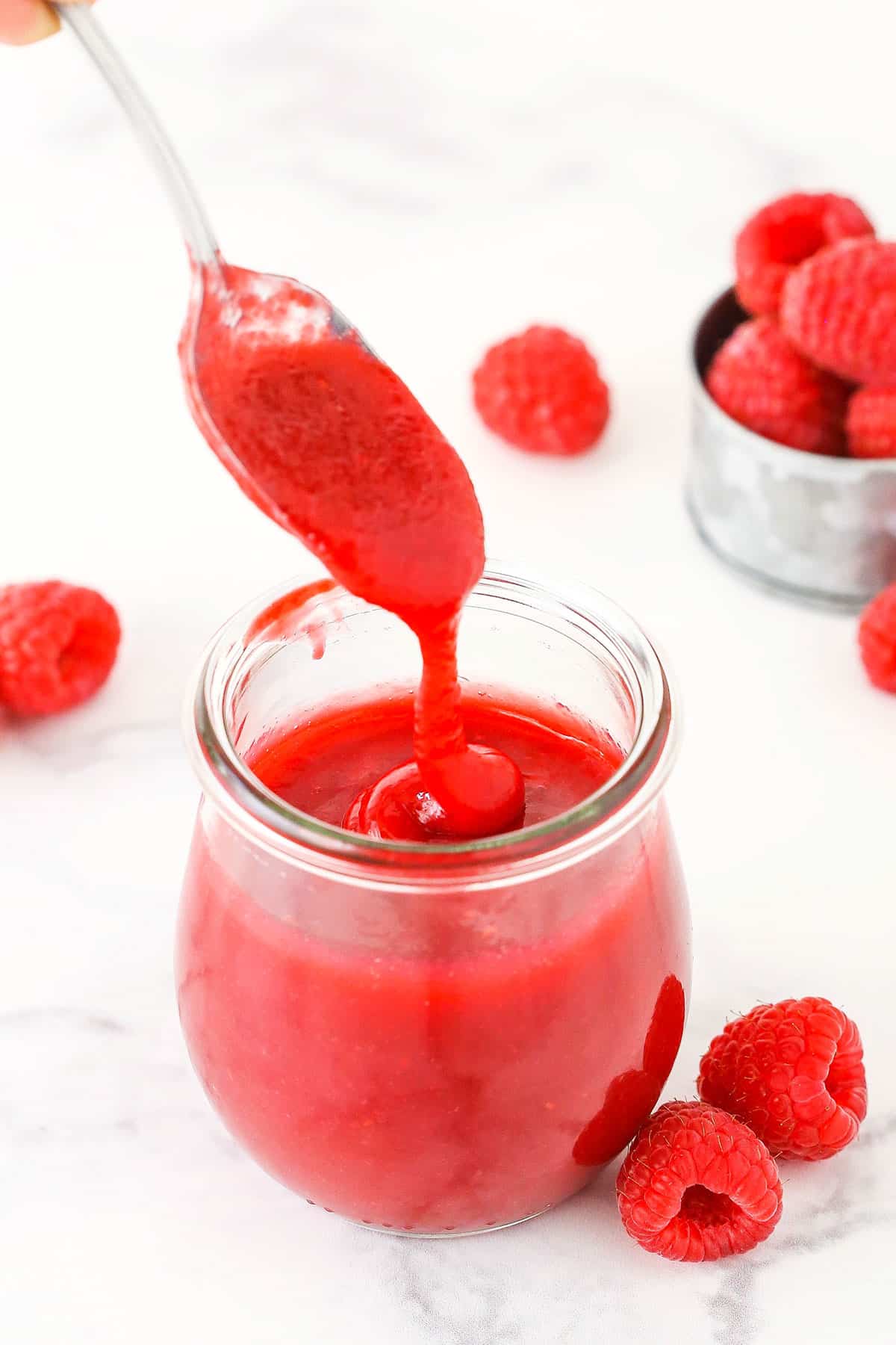 Raspberry sauce dripping off a spoon into a clear glass jar with raspberries in the background