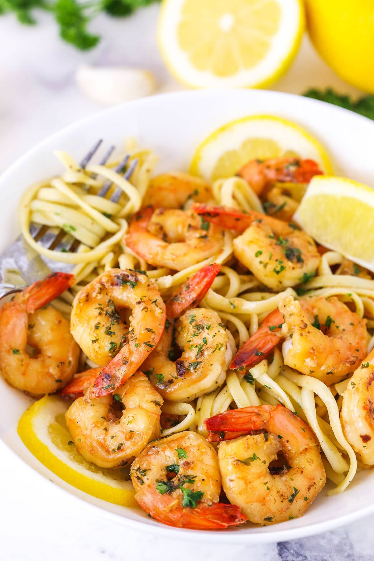 Garlic Butter Shrimp Scampi over linguini noodles with cut lemons in a white bowl with a fork