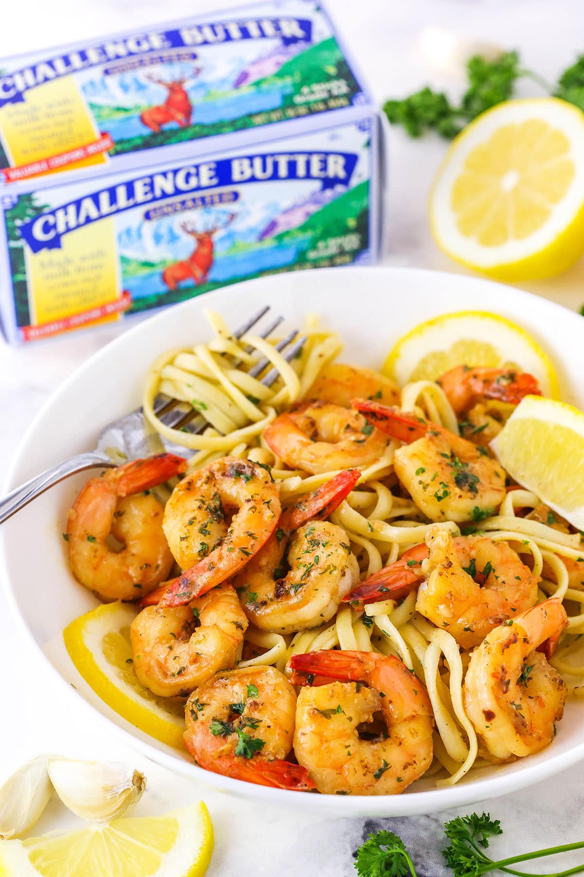 Garlic Butter Shrimp Scampi over linguini noodles with cut lemons in a white bowl with a fork and Challenge Butter in the background