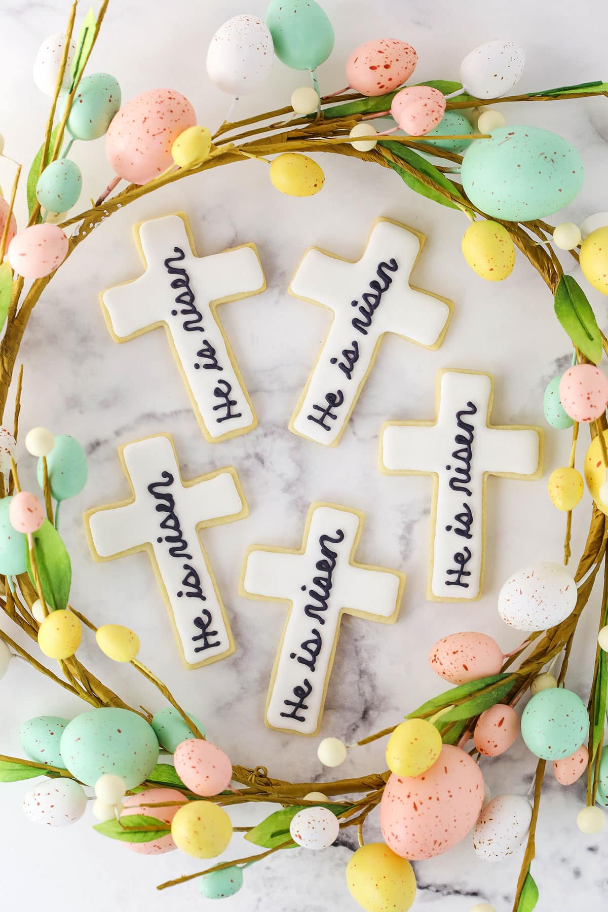 Five "He Is Risen" Cutout Cookies on a white marble table with an Easter wreath around them