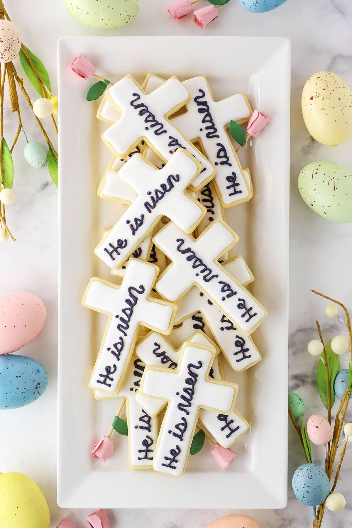 Overhead view of "He Is Risen" Cutout Cookies layered on a white platter with painted Easter eggs in the background