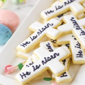 "He Is Risen" Cutout Cookies layered on a white platter with painted Easter eggs in the background