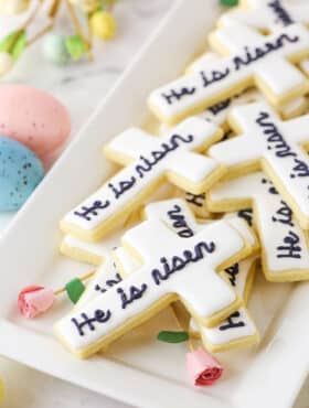 "He Is Risen" Cutout Cookies layered on a white platter with painted Easter eggs in the background