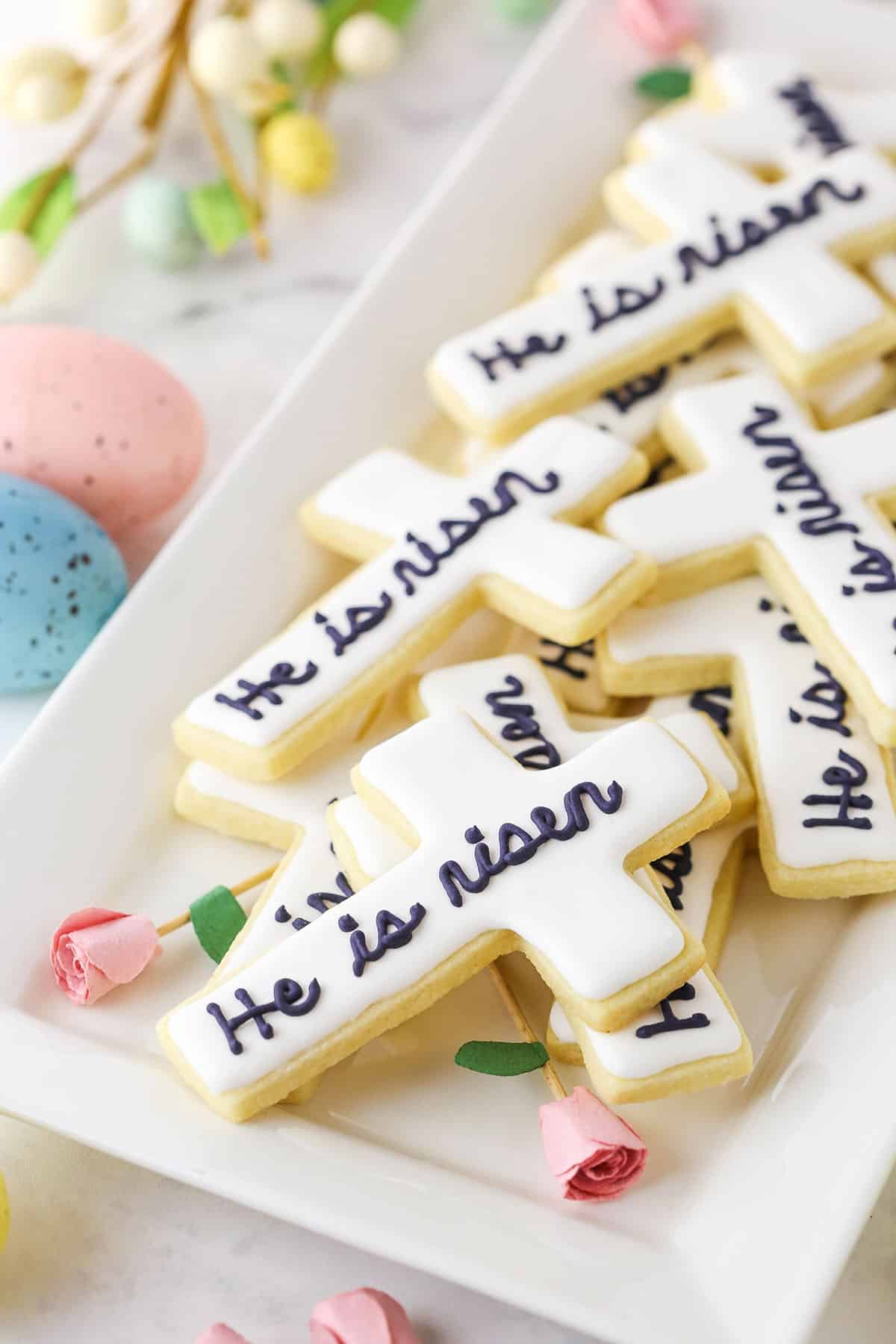 "He Is Risen" Cutout Cookies layered on a white platter with painted Easter eggs in the background
